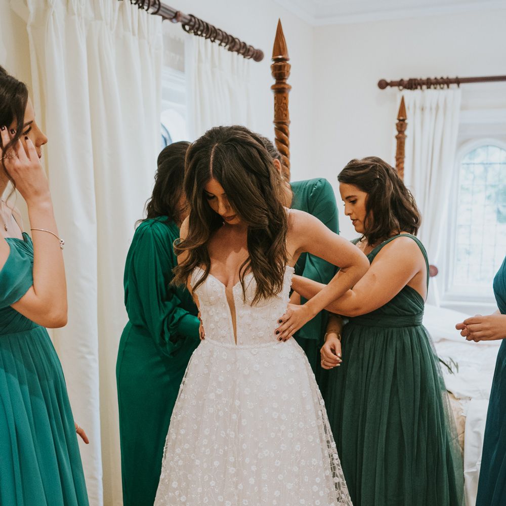 Bride in cami strap Pronovias wedding dress with mesh applique skirt has back of her dress tied by bridesmaids in mixed green bridesmaid dresses before Notley Abbey wedding