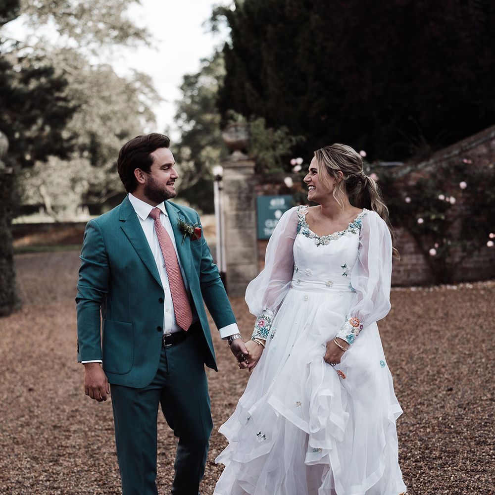 Groom in blue suit and pink tie holding hands with bride in long mesh sleeve wedding dress and white trainers walks across drive at Iscoyd Park