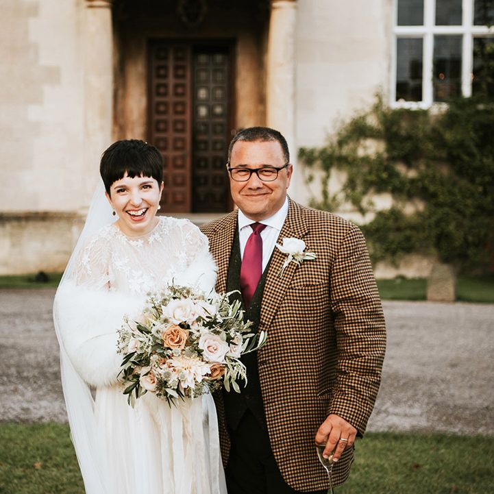 Bride with short hair in a white faux fur stole with her father in a check jacket outside Elmore Court 