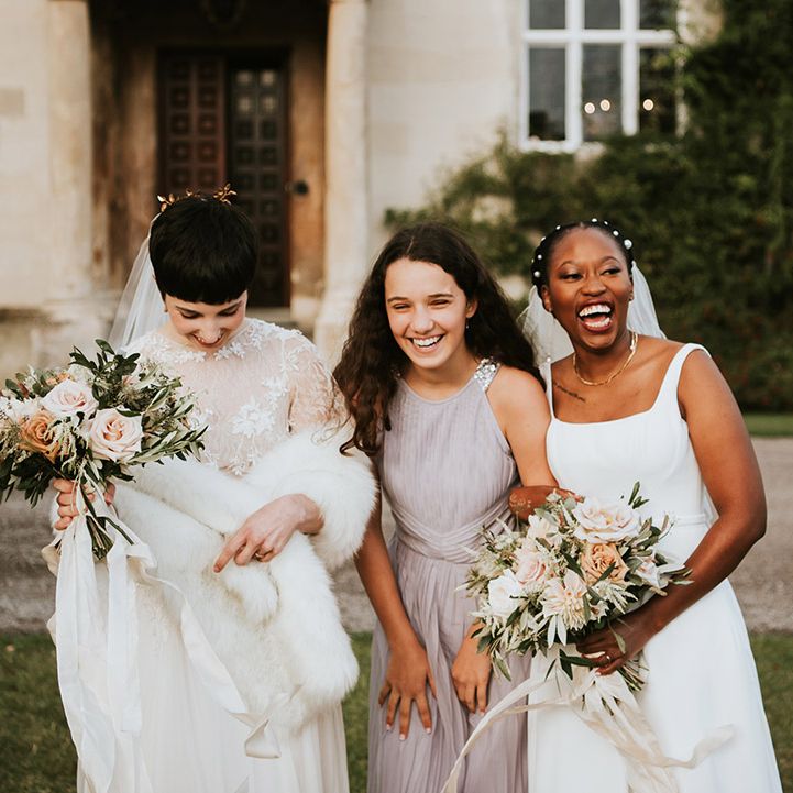 Two brides laughing with their junior bridesmaid in a dusky pink dress outside their country house wedding venue 