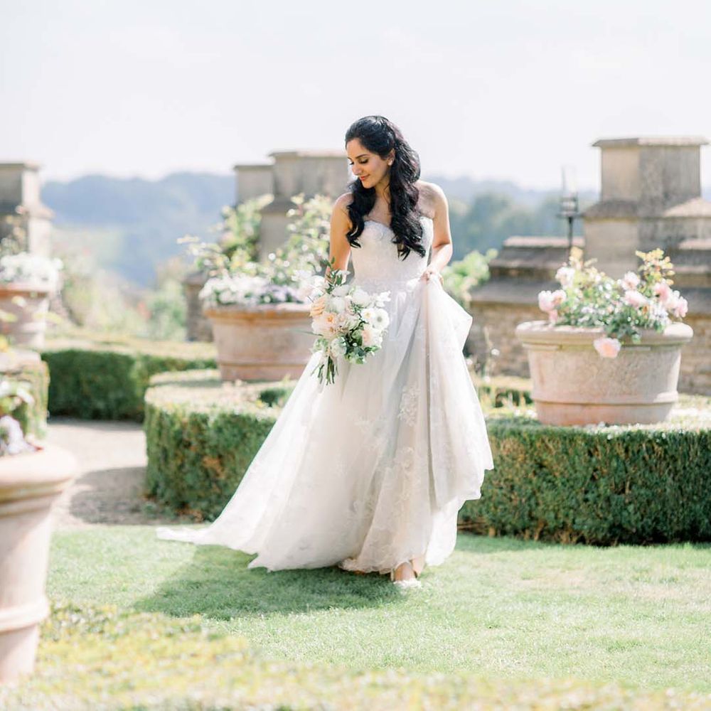 Indian bride holds her bridal gown outdoors on her wedding day whilst she holds floral bouquet 