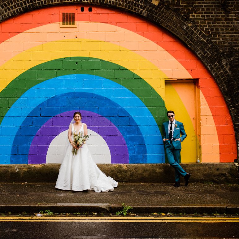 Groom in sunglasses and blue suit and bride in v neck wedding dress stand in front of rainbow backdrop at 100 Barrington