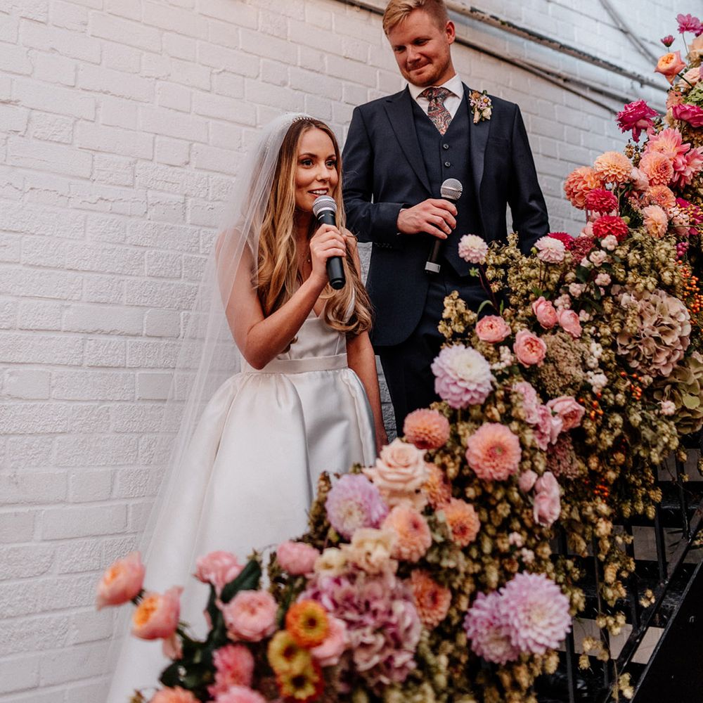 Bride and groom give a joint wedding speech at The Loft Studios on the staircase decorated with pink wedding flowers 