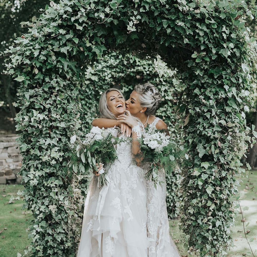 Couple at LGBTQI+ wedding holding white and green bouquets embracing under trees 