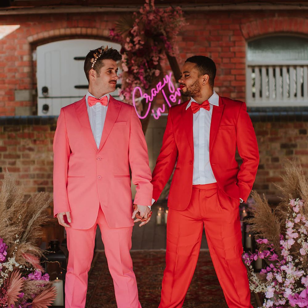 Groom in a red suit with winged eyeliner makeup holding hands with his partner in a pink suit, bow tie and gold crown