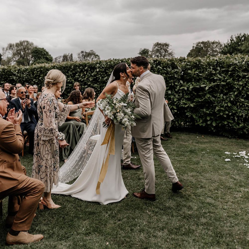 Indian bride in a satin wedding dress and pearl veil walking the aisle at her outdoor garden wedding ceremony 