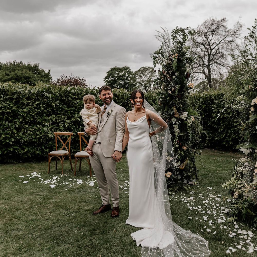 Bride in a minimalist slip wedding dress and bespoke pearl veil and groom in a beige suit at their outdoor garden wedding 