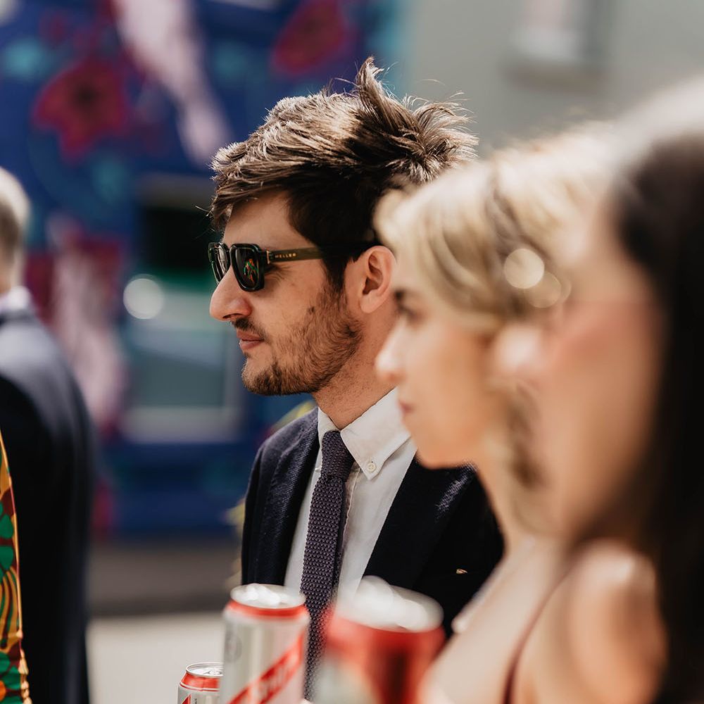 Wedding guests hold cans of beer as they stand outdoors in the sunshine 