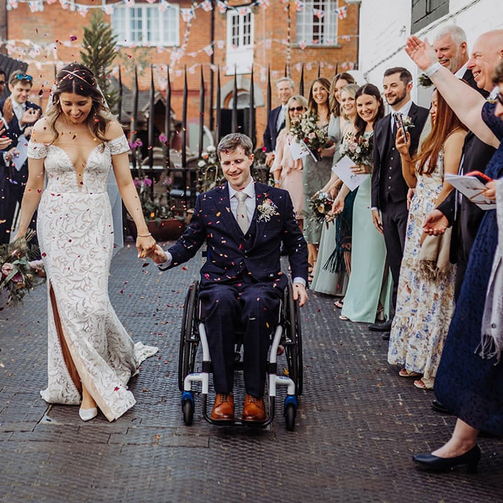 Bride in boho lace off the shoulder wedding dress with gold hair piece holding hands with the groom in a dark blue suit and gold tie as they have their confetti moment