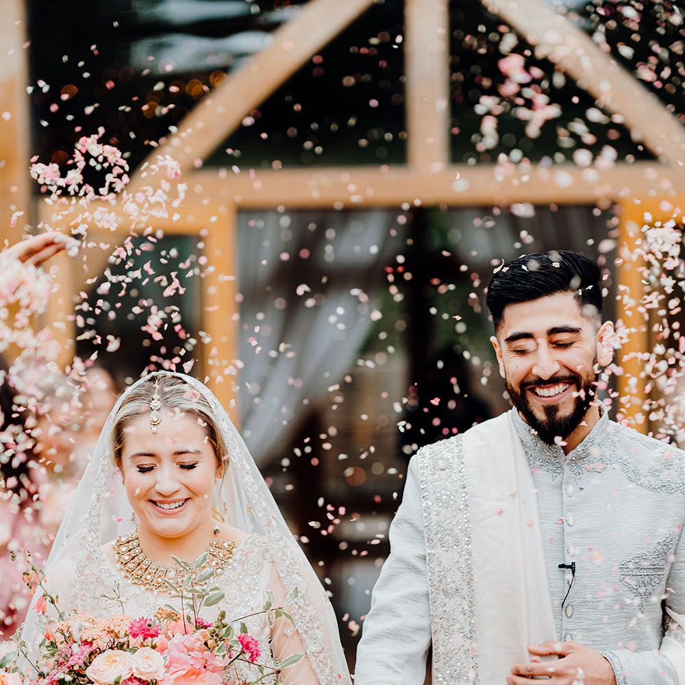 Bride holding bright floral bouquet and her groom walk through confetti outdoors at The Barn At Upcote after wedding ceremony