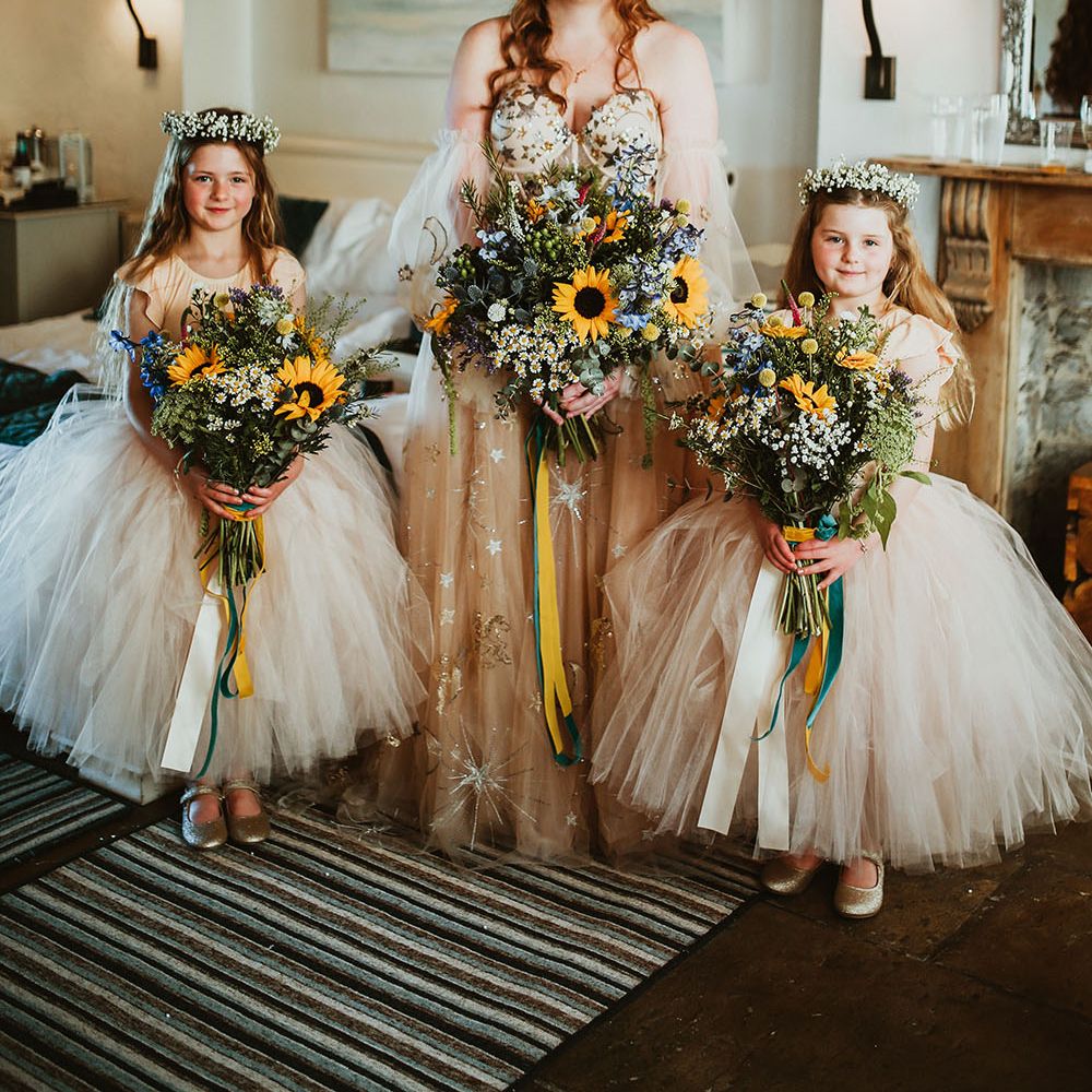 Bride wears pink starry wedding dress with sheer off the shoulder sleeves whilst stood with flower girls in pink ballerina dresses all holding sunflower bouquets