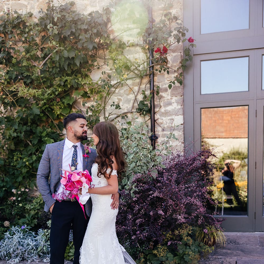 Bride & groom stand outdoors on their wedding day at the Priston Mill as bride holds bright pink floral bouquet 