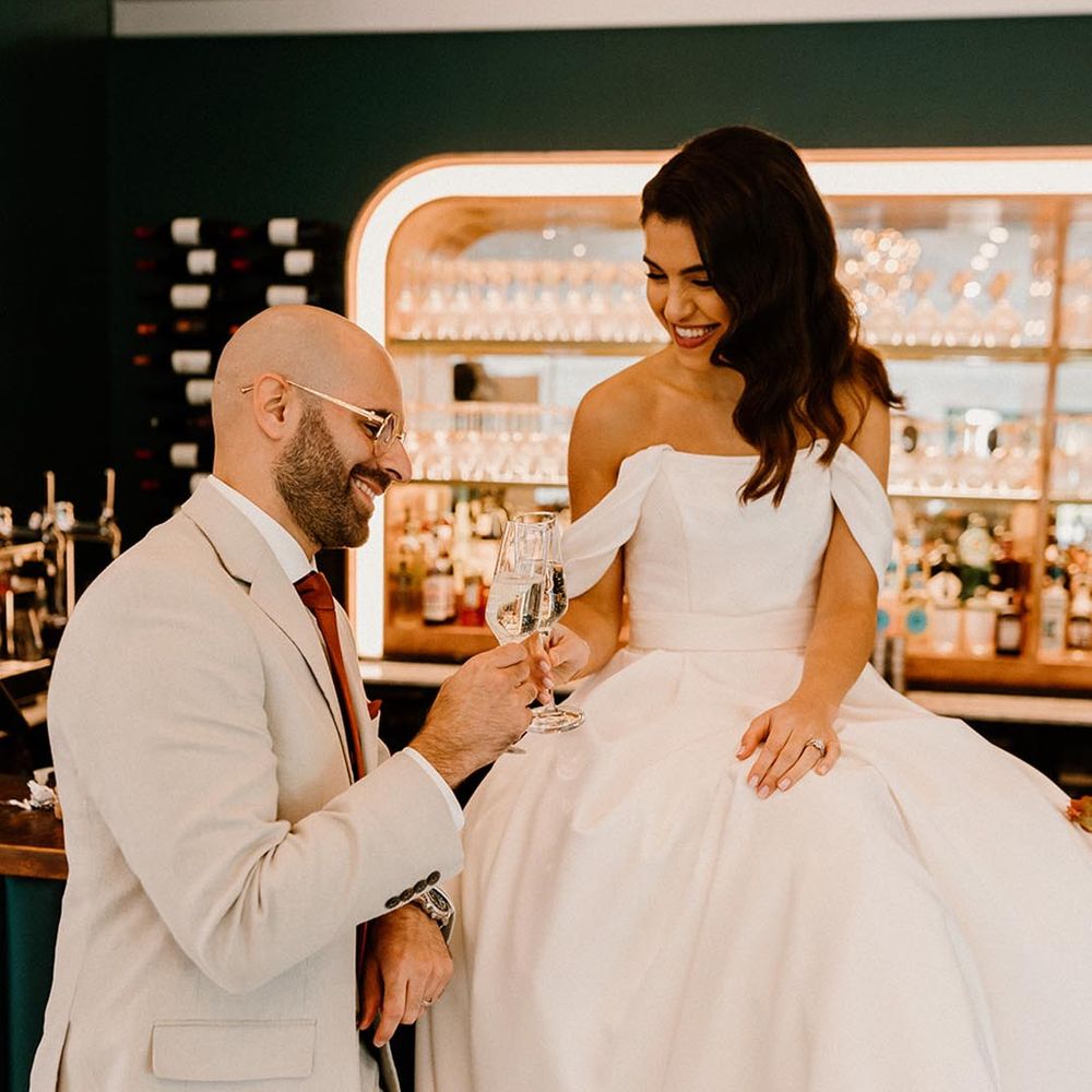 Bride in her off the shoulder wedding dress sits on the bar making a toast with the groom 