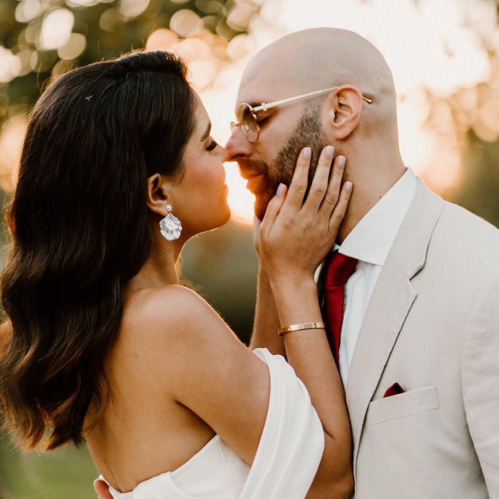 Bride with pearl earrings leaning in to kisses the groom in a pale grey suit and red tie