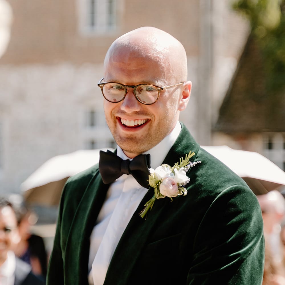 Groom wears black tie green velvet suit jacket and white floral buttonhole 