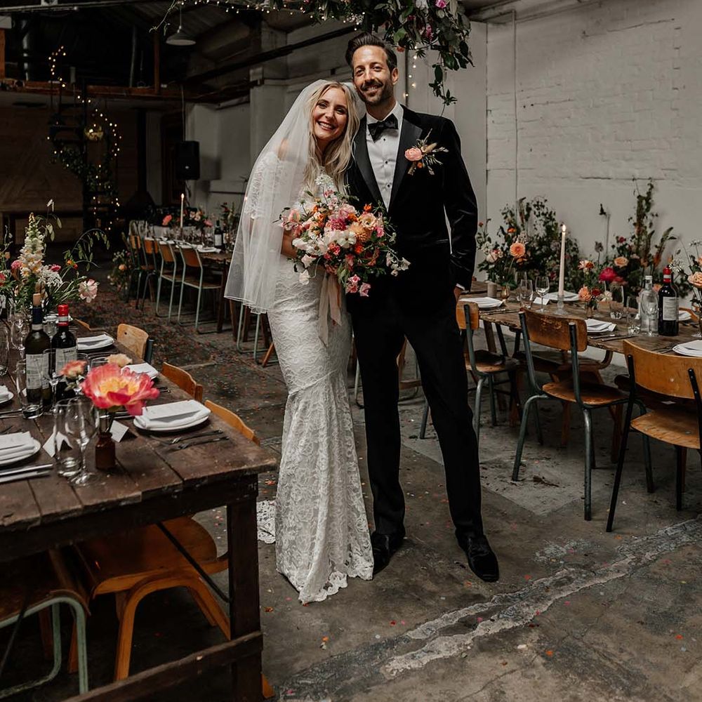 Bride in boho two piece lace bridal outfit with large colourful bouquet and groom in black tux with velvet look bow tie standing in reception room at Hackney Studios