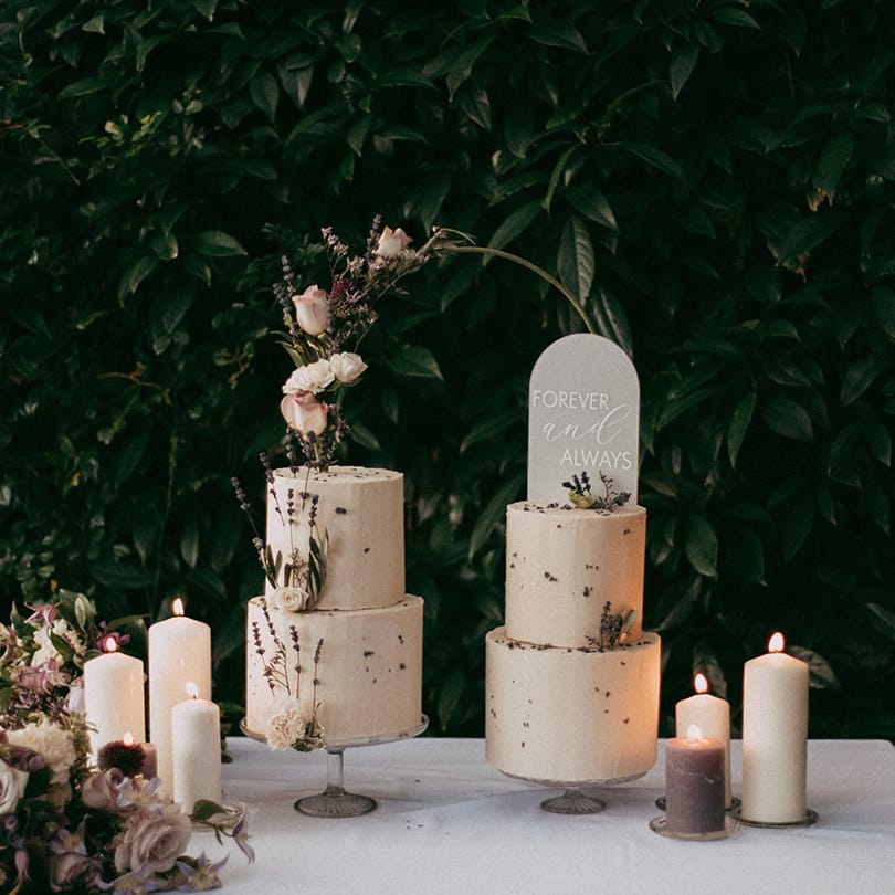 Two tiered buttercream wedding cakes with dried lavender and white rose decorations, white and lavender pillar candles, and neutral toned rose and foliage flower arrangements 