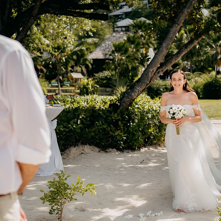 Bride in off the shoulder lace wedding dress with sweetheart neckline and lace floral details and cathedral length veil and tropical bouquet walking towards groom at Seychelles elopement 