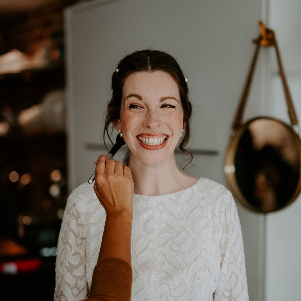 Bride in long sleeve lace vintage wedding dress and closed toe satin heels with pearl earrings and pearl hair clips getting makeup done before autumnal wedding
