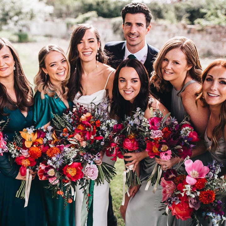 Bridesmaids in mismatched green bridesmaid dresses stand smiling with the bride and groom all holding bright pink, red, and orange wedding bouquets 