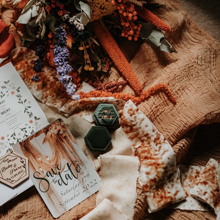 Boho wedding stationery on orange fabric with forest green velvet ring holder, golden wedding rings and colourful dried flowers