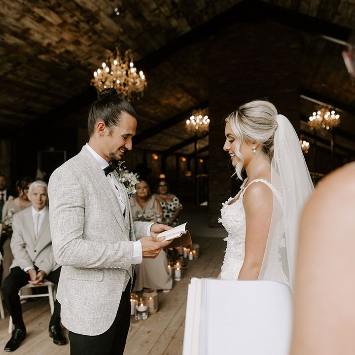 The groom in light grey suit stands facing the bride at the altar reading out his personalised wedding vows from his custom booklet 