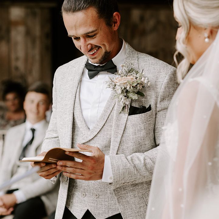 Groom in light grey three piece suit with dark bow tie and pocket square reading out personalised vows for the handfasting ceremony 