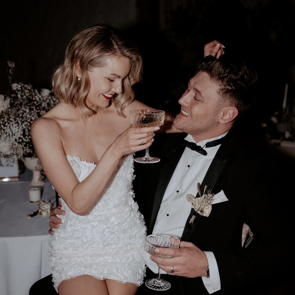 Bride in short wedding reception dress sitting on the groom's lap in a black tuxedo drinking champagne 