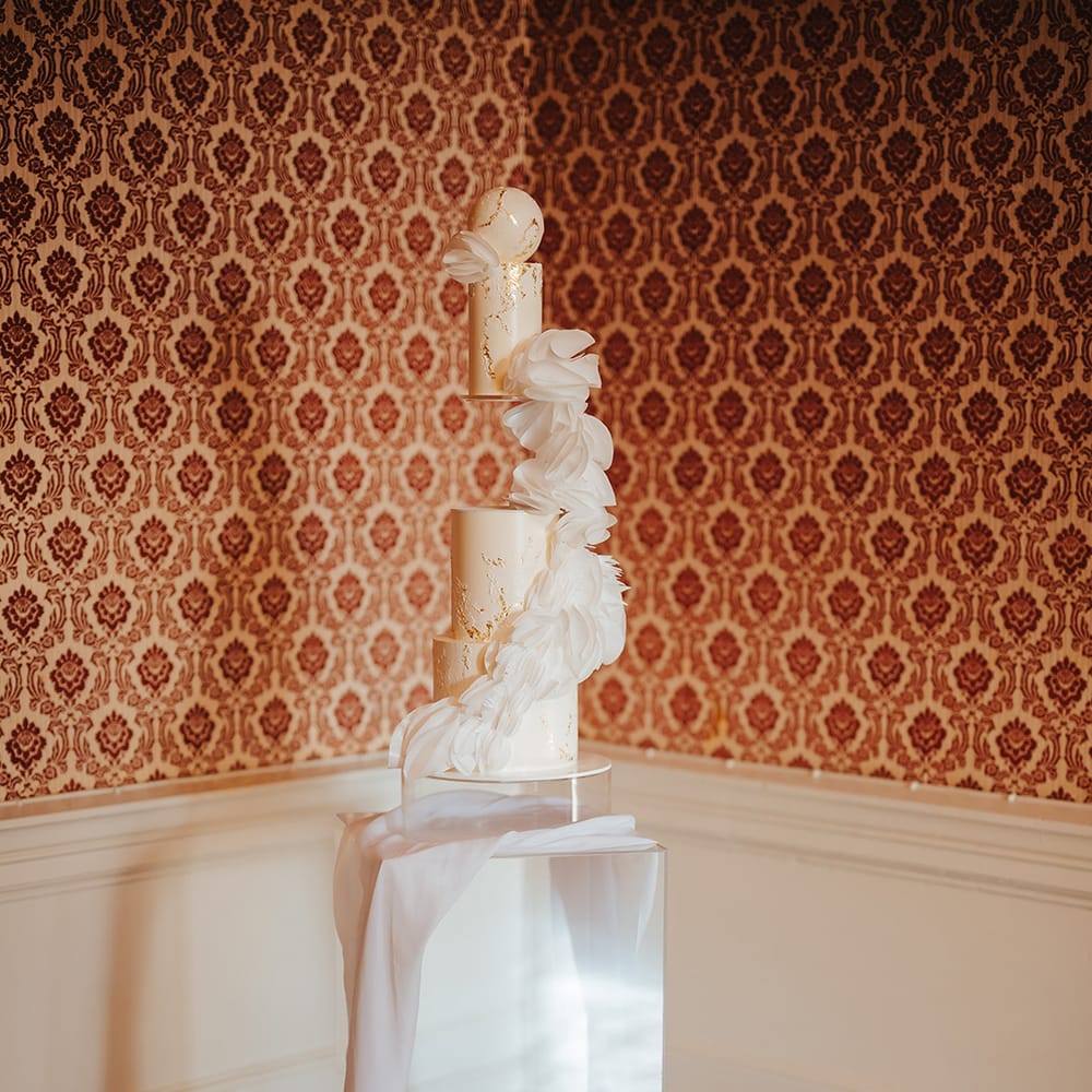 Artistic wedding cake - three tiered wedding cake with white feather like decorations, white cake ball on the top and white wedding drapes on large clear cake stand 