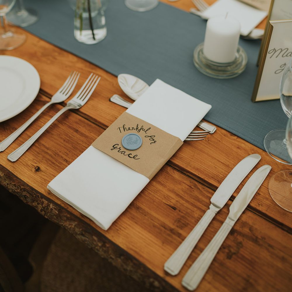 Wooden table with silver cutlery and blue table runner decorated with bud vases of daisies and a white napkin with cardboard holder and wax seal
