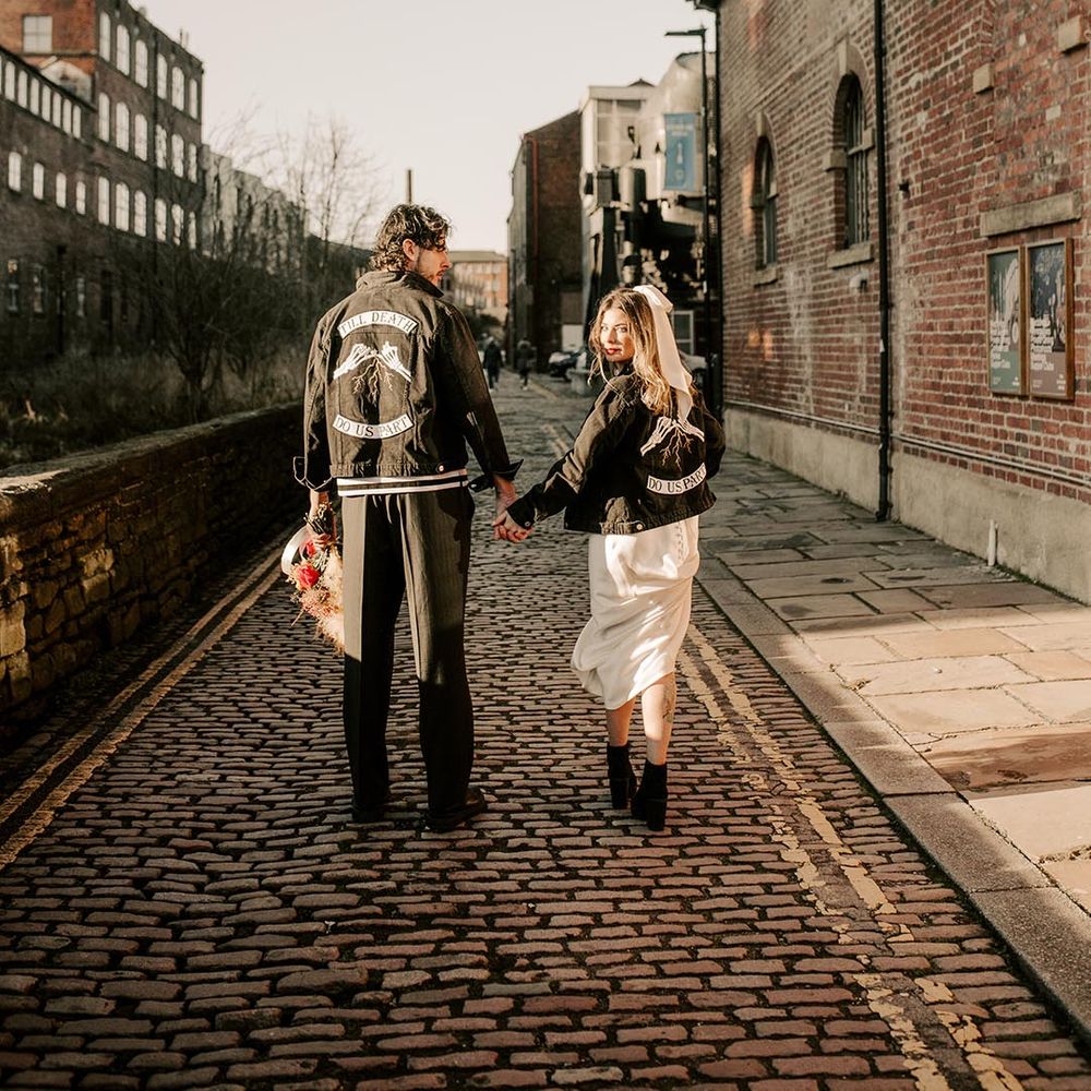 The bride and groom wear matching his and hers black denim jackets with skeleton hands making a pink promise and writing reading 'till death do us part'