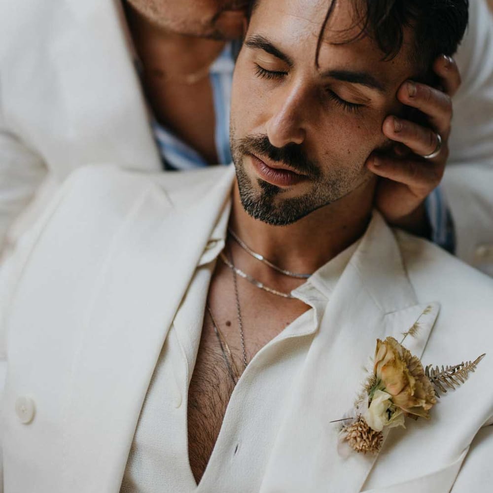 Groom in white blazer, dusky orange garden rose and dried flower boutonniere, white shirt and silver jewellery with groom in white suit and white and blue shirt 
