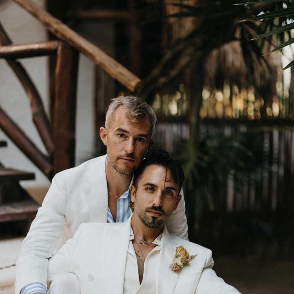 Groom in mens white wedding suit with dusky orange garden rose and dried flower boutonniere, white shirt, white suit trousers and silver jewellery posing with groom in white blazer, light blue and white striped shirt, dusky orange garden rose and dried flower boutonniere, white suit trousers and gold jewellery 