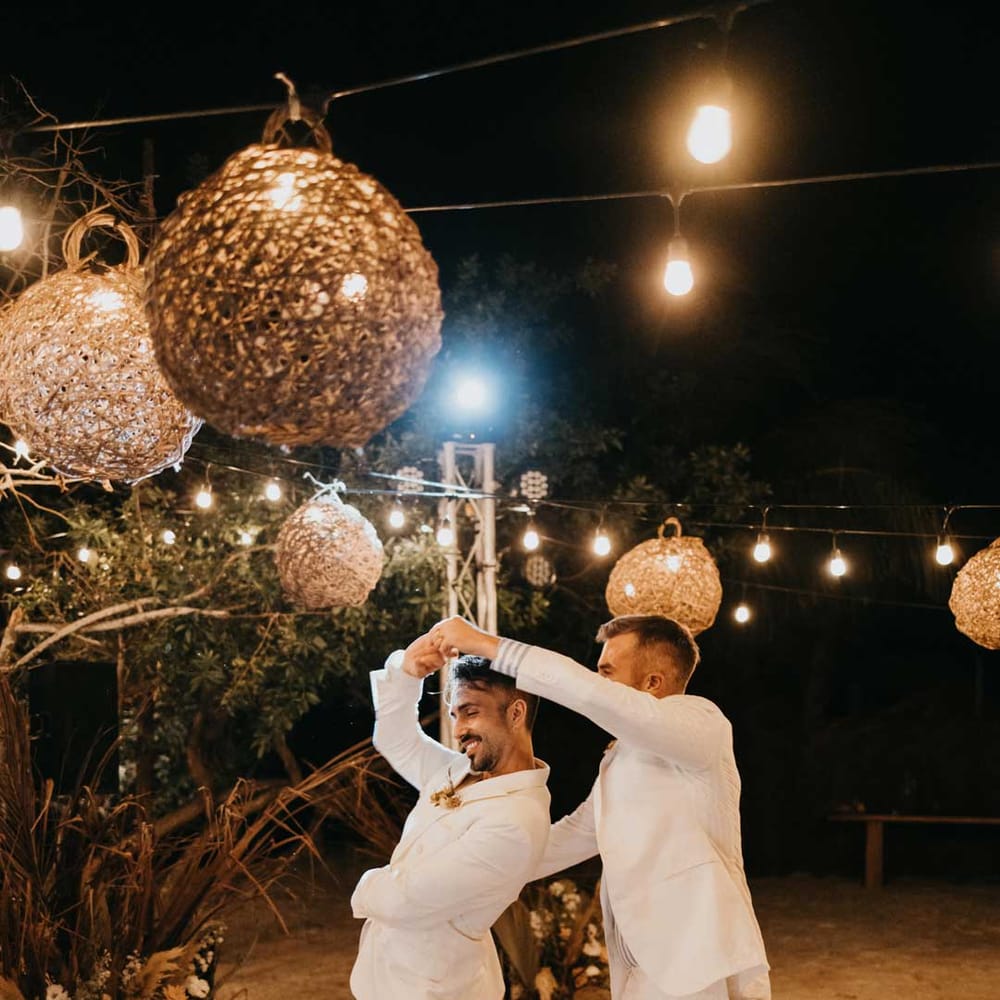 Grooms in matching white grooms suits dancing on DIY hand painted wedding dance floor under twine lampshades and fairy lights 
