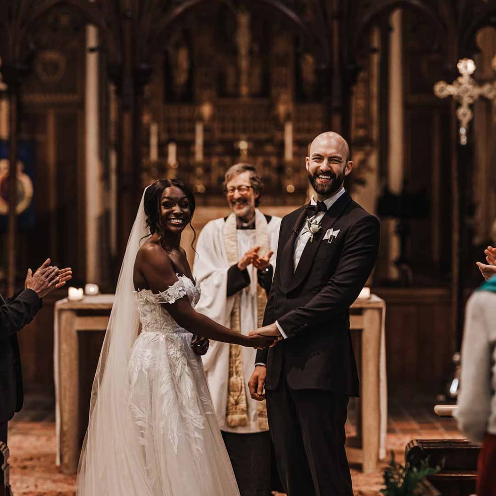 Groom in classic black three piece grooms suit with bow tie and white rose and foliage boutonniere in church holding hands with bride in sweetheart neckline lace wedding dress with detachable off the shoulder sleeves in church wedding 