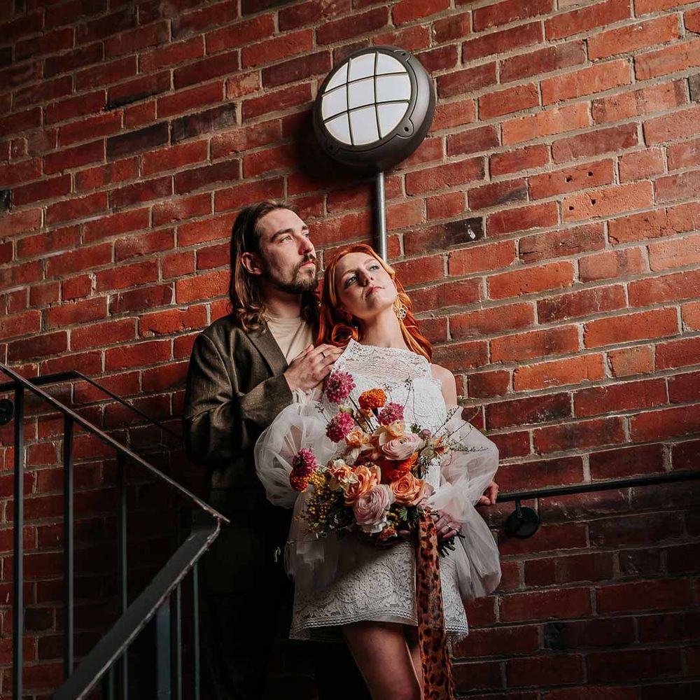 Bride in 70s inspired boatneck short lace wedding dress with detachable tulle puff sleeves and large platform gold wedding heels holding large colourful mixed flower bridal bouquet with groom in brown blazer, white shirt, grey suit trousers and vans shoes 