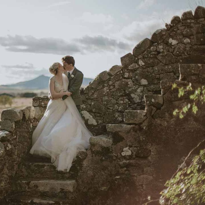 Bride in dramatic sleeveless princess wedding dress kissing groom in classic grey grooms suit on staircase at Barra Castle wedding venue Scotland