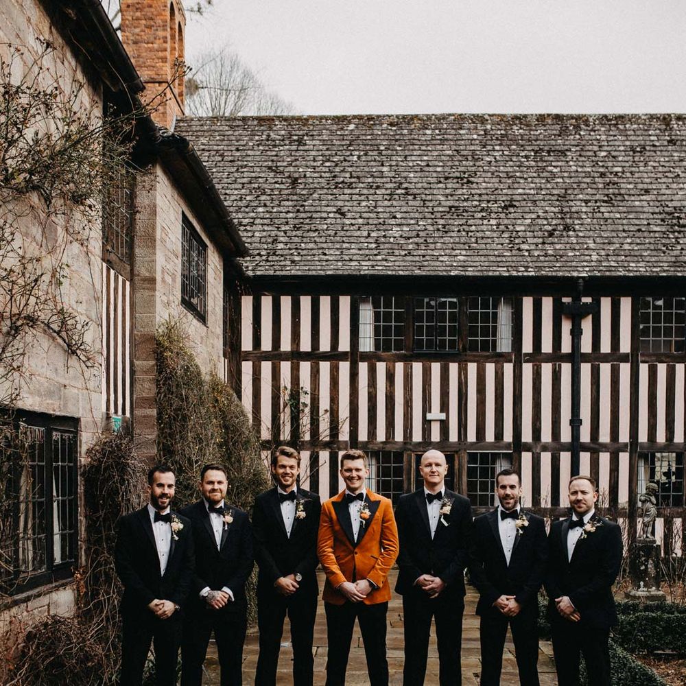 Groomsmen wearing classic black grooms suits with bowties standing with groom in crushed burnt orange velvet grooms blazer, black bowtie and black suit trousers outside Brinsop Court wedding venue