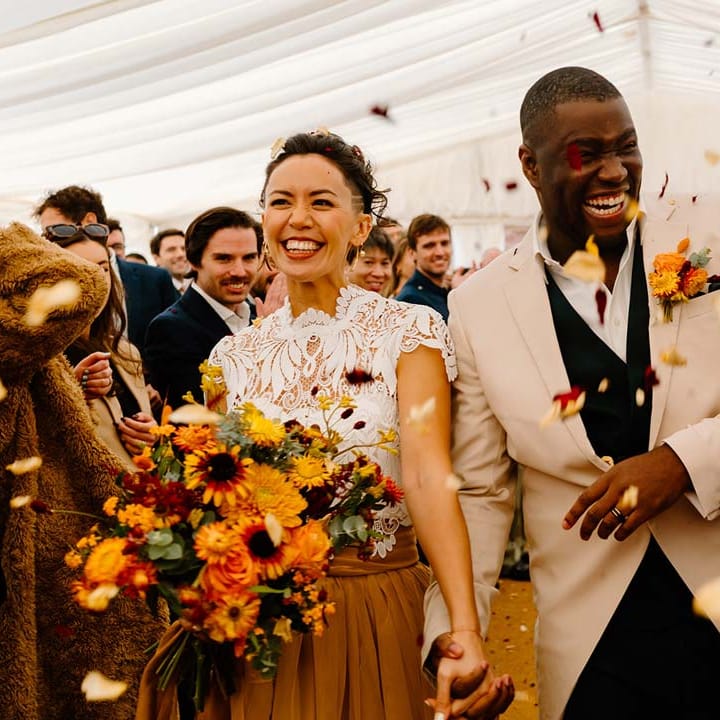 Groom in cream morning suit with forest green grooms waistcoat and trousers, forest green pocket square and orange boutonniere doing colourful dried petal confetti wedding exit at Broadfield Court hand in hand with bride in white mesh lace bridal crop top and yellow wedding skirt