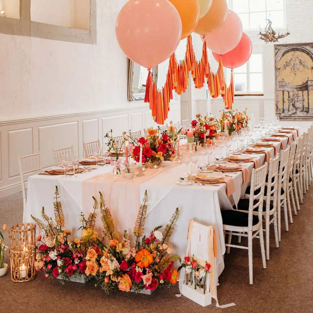 Wedding tablescape with white wedding tablecloth, burlap place mats, tapered candles, pink and orange wedding stationery, and floral wedding table centrepieces with pink and orange garden roses, orange floribunda, yellow ranunculus flowers, foliage and Iceland poppies with red, pink orange and yellow balloons, wedding streamers and fairy lights at Merriscourt wedding venue