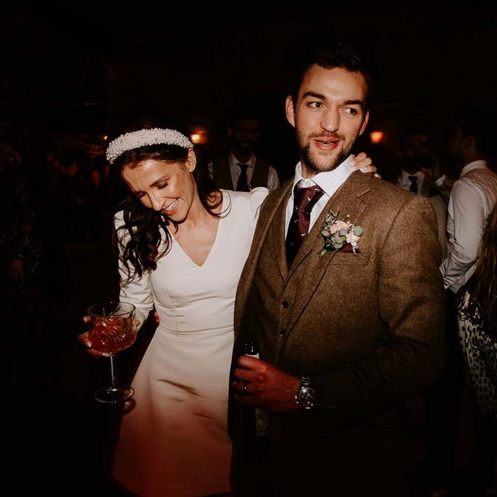 Bride in long sleeve short wedding dress and pearl bridal headband posing with groom in three piece chocolate brown grooms suit 