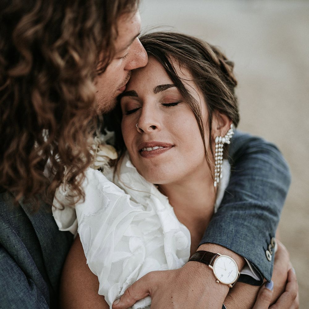 Bride with a nose ring and chandelier earrings in a ruffle sleeve wedding dress being kissed on the head by her curly hair groom
