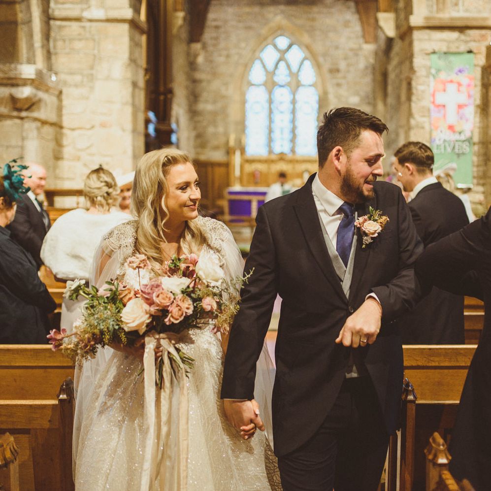 Bride and groom walking up the aisle as husband and white elbow bumping guests 