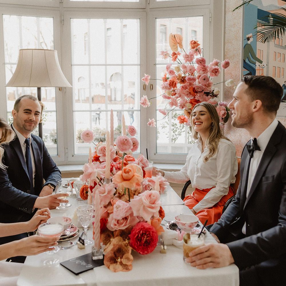 Bride, groom and two guests seated at the wedding table with cocktails and peach wedding flower centrepieces including pink, peach and burgundy ranunculi, garden roses and orchids