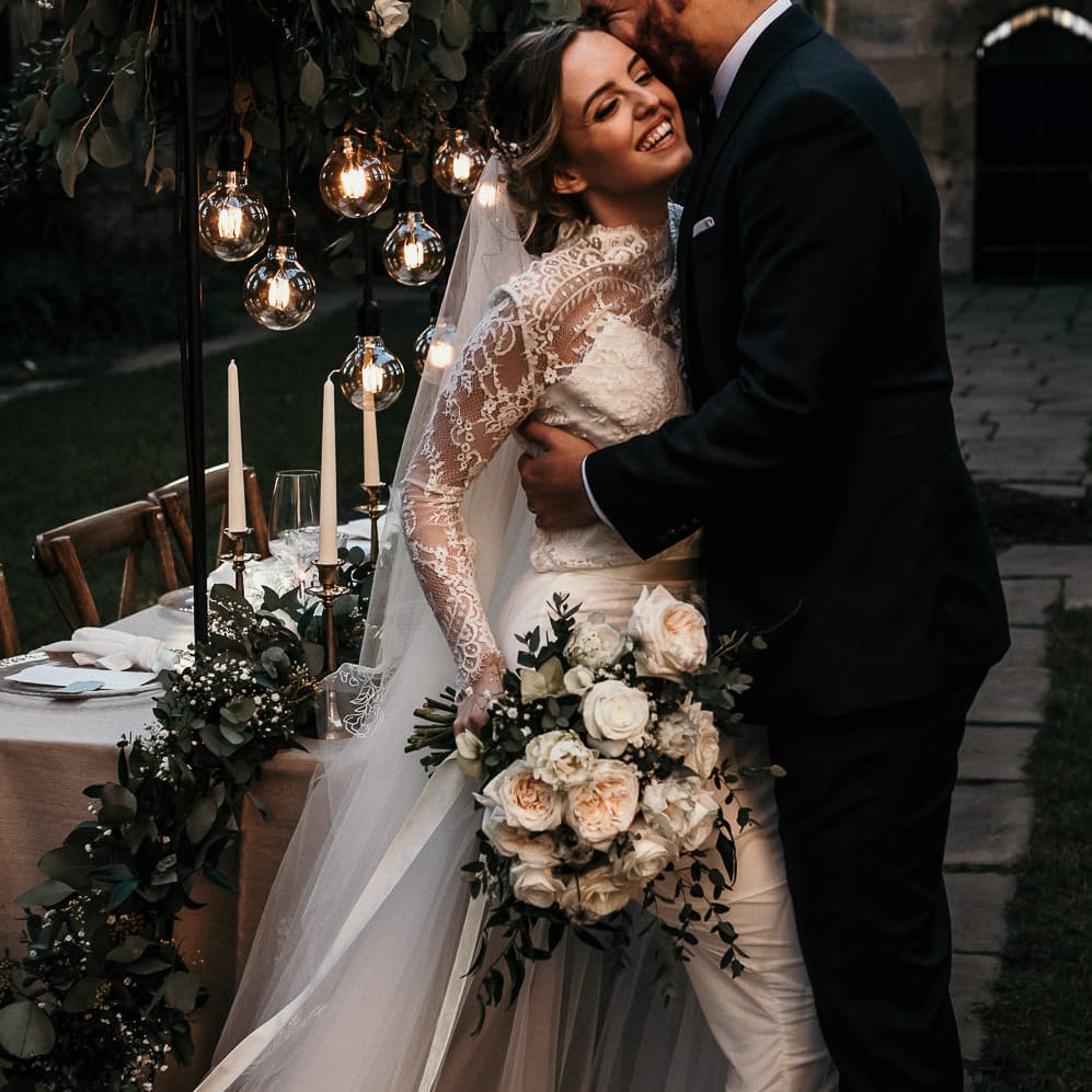 Groom in a black suit laughing with his bride in a long sleeve lace wedding dress as they sit on the edge of their elegant tablescape with festoon lights, greenery table runner and candles 