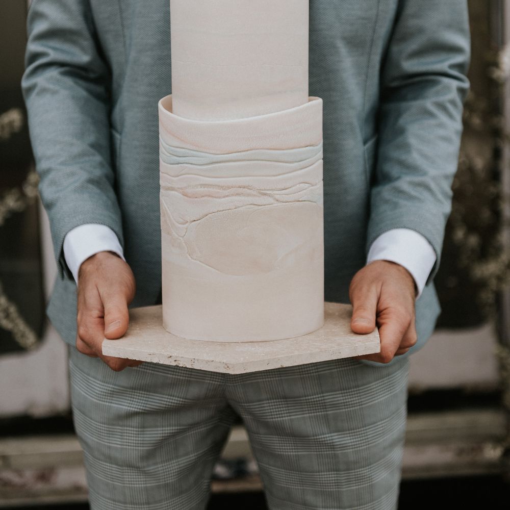 Groom holds wedding cake whilst wearing pastel blue suit 