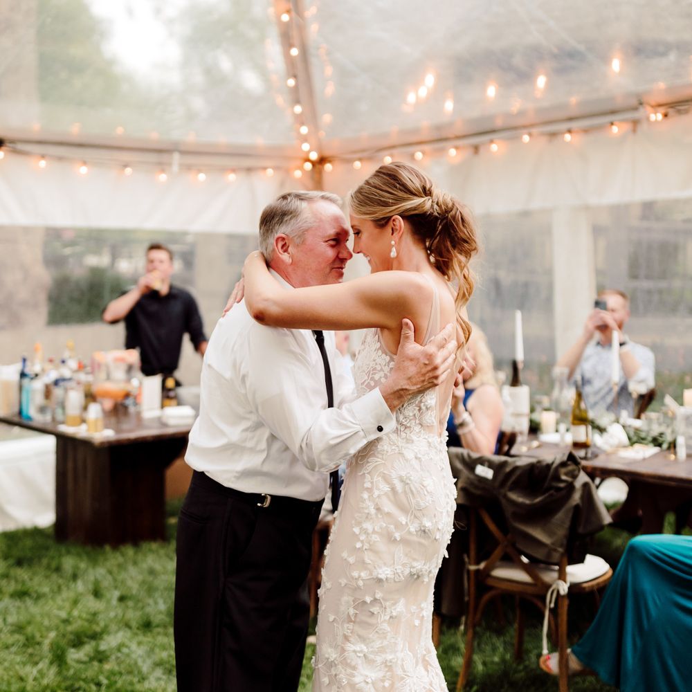 Bride with curled ponytail in white embroidered Made With Love wedding dress dances with arms around man in white shirt in fairy lit marquee at backyard wedding