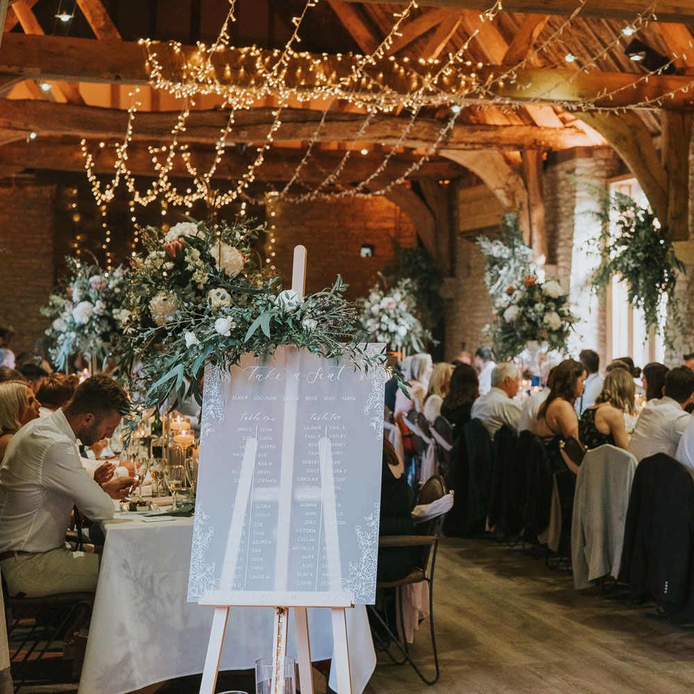 Guests sit at long white wedding tables in stone room with wooden beams lit by string fairy lights with frosted acrylic table seating plan wedding sign at Tythe Barn wedding with barn wedding flowers