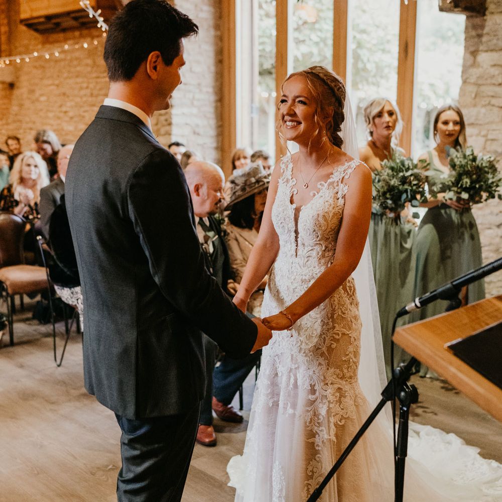 Bride in lace and tulle wedding dress stands holding hands with groom in dark suit during barn wedding ceremony