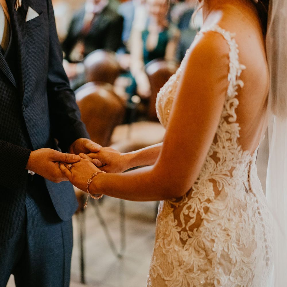 Bride in open back lace wedding dress and veil wearing bracelet holds hands with groom in dark three piece suit during wedding ceremony